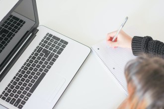 foto de um computador e do lado aparece uma mão segurando um lápis e escrevendo num caderno