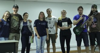 Fotografia em médio plano. 8 pessoas entre homens e mulheres lado a lado olhando para a câmera e sorrindo. Eles estão em cima de um palco.