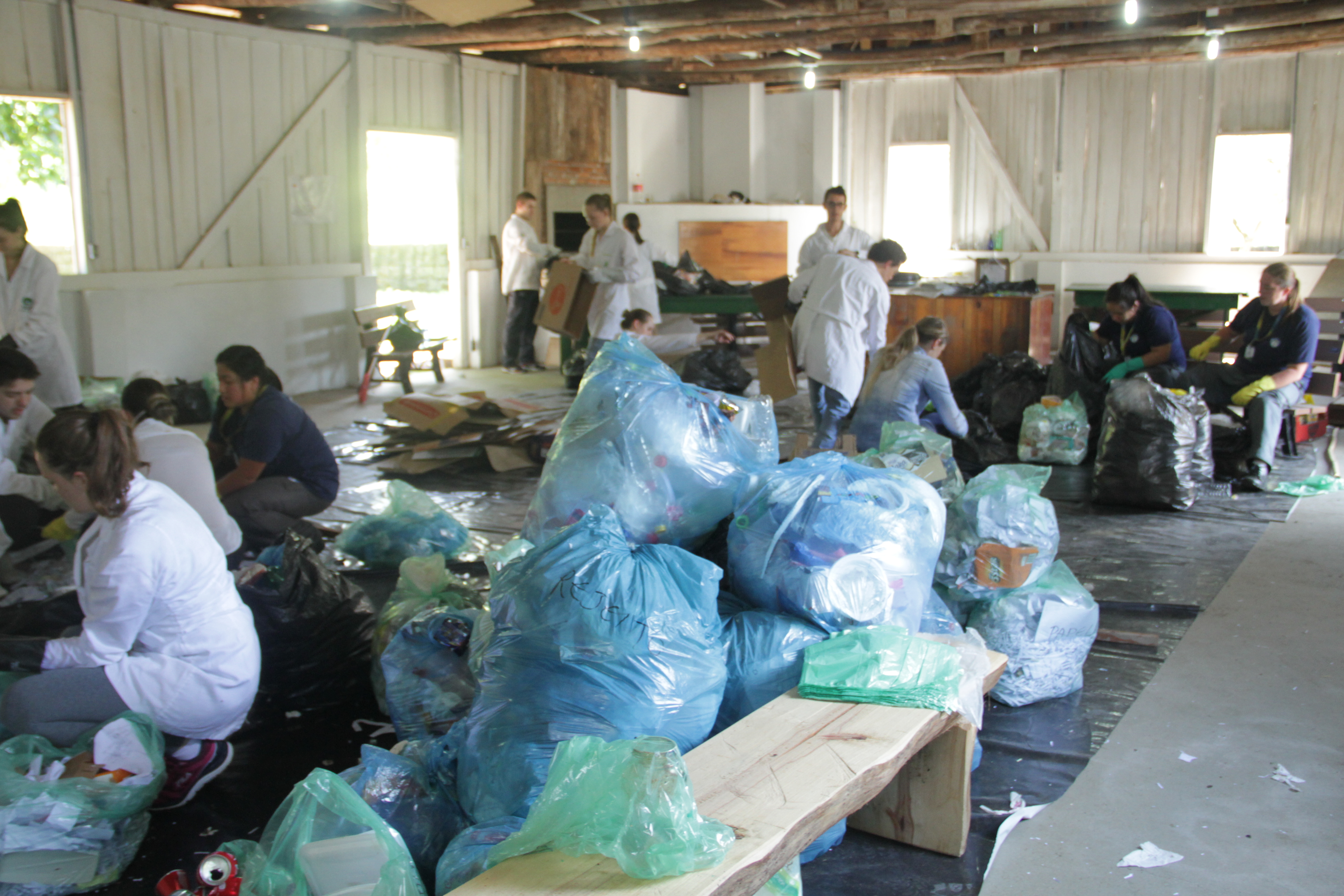 Foto em plano aberto, porém de outro ângulo, mostrando os sacos de lixo, caixas de papelão e a equipe trabalhando.