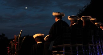 Foto de formatura: várias pessoas sentadas de costas para a máquina, vestidas com beca e capelo. Acima deles a lua cheia. O evento é ao ar livre.