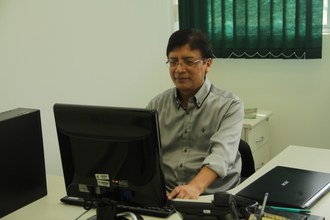 Fotografia em médio plano, com um homem sentado de frente para uma tela de computador. O homem está de frente para a câmera. No fundo, uma cortina persiana, na cor verde.