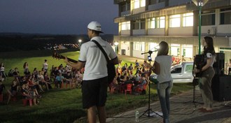 Foto noturna em um espaço aberto com três artistas se apresentando, em primeiro plano, e a plateia assistindo (capa).