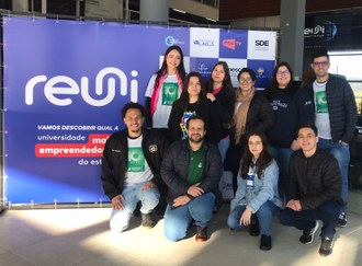 Foto de jovens em pé e abaixados ao lado de painel com a marca do desafio Reuni