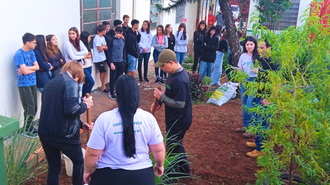 Foto do pátio da escola com vários estudantes encostados na parede da escola e graduandos conversando e trabalhando na terra