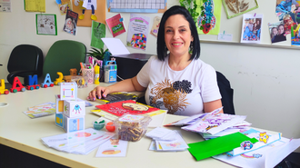 Foto da professora, uma mulher branca, de cabelos curtos e escuros. Ela está sorrindo, sentada atrás de uma mesa. Sobre a mesa estão vários materiais utilizados por ela em sua atuação para ensinar futuros professores a alfabetizarem crianças