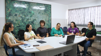 Imagem de seis pessoas, sendo quatro mulheres e dois homens. Eles estão em uma sala de reuniões, sentados ao redor de uma mesa oval, com um notebook à frente