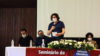 Foto de parte de uma mesa de honra, com uma pessoa falando ao microfone e as demais, sentadas atrás da mesa