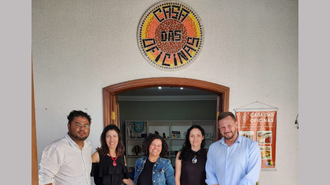 Dois homens e três mulheres estão em frente à porta de uma edificação. Acima da porta há um mosaico colorido com o texto "Casa das Oficinas"