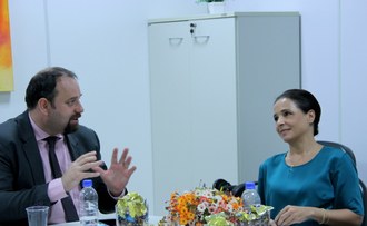 Imagem de um homem à esquerda, e uma mulher à direita. Estão sentados à mesa. Ambos conversam e gesticulam