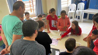 Imagem de dois adultos em frente a uma mesa, observando o que é mostrado pelas crianças que estão ao redor da mesa.