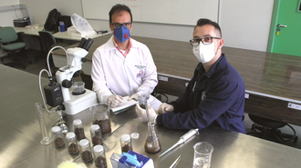 foto em que aparecem duas pessoas paramentadas com EPIs, em um laboratório, sentados, em frente a uma bancada. Na bancada há vidrarias e um microscópio