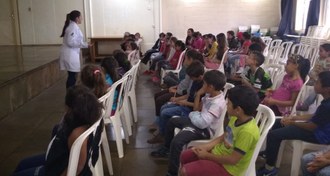 Estudante de jaleco fala, de frente para as crianças, que estão sentadas em cadeiras brancas, em uma sala