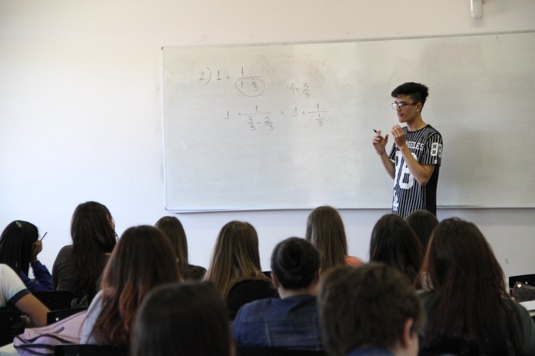 Estudante explicando resolução no quadro branco. Jovens das escolas aparecem de costas, olhando para o graduando