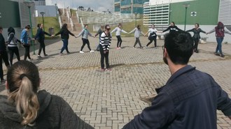 No campus, em uma calçada em frente aos prédios de laboratórios, estudantes estão em formação de círculo, de mãos dadas, e uma das estudantes está no centro