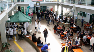 Imagem do alto do hall da UFFS - Campus Chapecó com muitas pessoas circulando, uma tenda com vários jovens de jaleco e outras sentadas em cadeiras ao redor de mesas da cantina