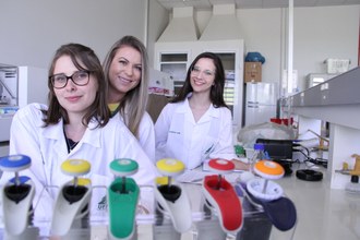 No laboratório, em frente à bancada, com ferramentas de laboratório em primeiro plano, Marta, Andréia e Maria Luiza aparecem olhando para a câmera, uma atrás da outra.