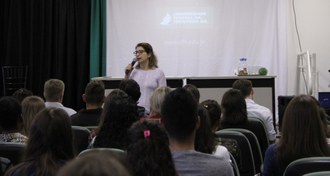Em um auditório, pessoas de costas, sentadas, observam uma mulher, de frente, falando ao microfone. Ao fundo, projetado na parede, o logo da UFFS - Campus Chapecó.