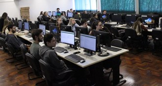 Foto em plano aberto mostra duas filas de computadores, com mesas dispostas de frente, todas com computadores. Estudantes estão sentados em várias cadeiras