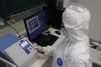 Na imagem aparece um equipamento de laboratório, um computador ao lado e uma pessoa mexendo no equipamento