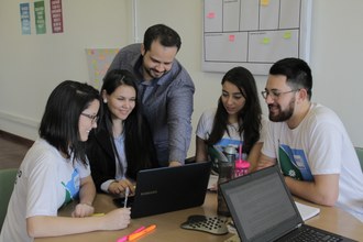 Três jovens mulheres e um homem estão sentados ao redor de uma mesa redonda e o professor está em pé, logo atrás deles, apontando para a tela de um notebook