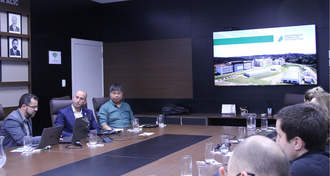 Foto em plano aberto mostra parte de uma mesa de reuniões, com pessoas sentadas ao redor, e, em frente, uma apresentação projetada na tela, com a foto da UFFS - Chapecó