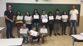 Na foto, registrada dentro de uma sala de aula, sete estudantes estão em pé e outros três estão agachados, todos seguram em suas mãos os certificados de participação no projeto. Ao lado deles estão Ronaldo José Seramim, um dos Coordenadores do Projeto e o professor Diogenes Marcante Ribeiro, Diretor do Colégio Estadual Vila Industrial.