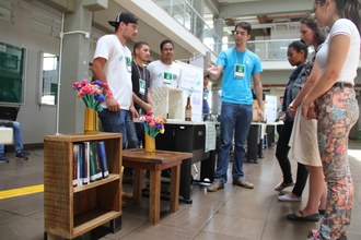Na imagem quatro estudantes realizam a apresentação do projeto de confecção e venda de móveis e decoração a partir de materiais reciclados. Ao lado dos estudantes uma estante baixa e uma mesa de centro feita de madeiras reaproveitada.