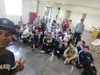 Um estudante segurando o celular faz uma selfie. Ao fundo um grupo de pessoas está posando para foto.