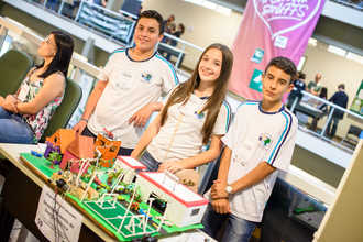 Três adolescentes estão em pé estão olhando para a câmera. A maquete do trabalho que estão apresentando está em frente aos adolescentes.