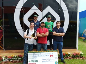 Na imagem os cinco integrantes da equipe posam com um vale em tamanho gigante, contendo a descrição do prêmio recebido. Um estudante segura nas mãos o troféu da equipe.