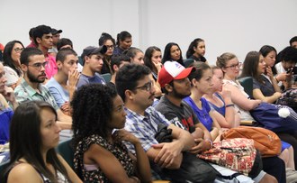 Na imagem, vários estudantes estão sentados em cadeiras enfileiradas observando algo a sua frente.