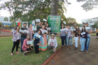 Na imagem várias pessoas estão em uma praça. Elas estão em pé e algumas seguram em suas mãos plaquinhas com hashtags com diversos dizeres. Atrás das pessoas tem uma árvore e nela está pendurado um banner com o logo da UFFS e nome dos cursos ofertados.