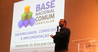 Na imagem o professor Élsio José Corá está no palco falando para o público. Ao fundo a projeção de imagem com título da palestra.