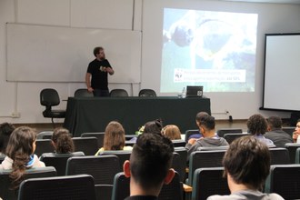 Estudantes sentados em auditório assistem apresentação de palestrante que está no palco. Ao lado esquerdo a imagem de um peixe é projetada no telão.