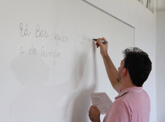Na imagem um professor está escrevendo no quadro branco e segurando uma anotação com a outra mão.