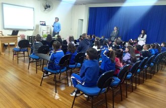 Dentro de uma sala de aula as crianças estão sentadas assistindo apresentação do professor.