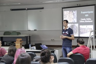 Na foto uma pessoa está em pé, em frente ao público. Ao fundo observa-se os slides que auxíliam na apresentação da palestra.