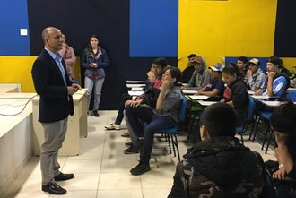 Na foto um homem está em pé falando para o público de jovens estudantes que permanecem sentados em um auditório.