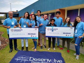 Na imagem, nove pessoas em pé estão posando para a foto. Algumas seguram em suas mãos as medalhas, cheques de premiação e troféu. A bandeira da Desenvolve está exposta no chão, na frente das pessoas fotografadas.