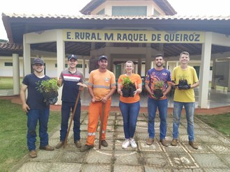 Na foto seis pessoas estão em pé em frente ao portal com o nome da escola. As pessoas seguram nas mãos as mudas e ferramentas de trabalho.