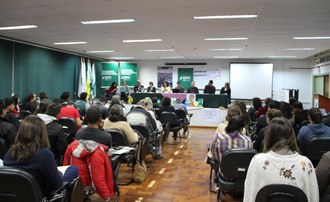 Na imagem a plateia sentada observa a mesa diretora em frente ao aduitório.