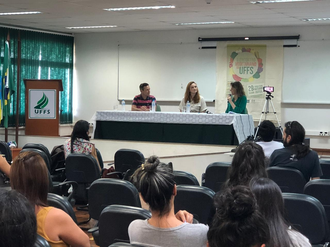 Na foto, o público no auditório acompanha a fala de três palestrantes que estão na mesa de honra.