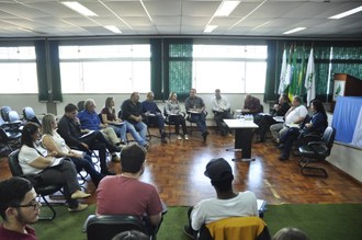 Na foto um grupo de pessoas está sentado em cadeiras em um auditório, formando um círculo.