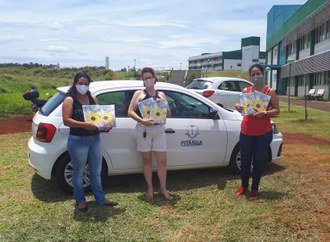 No centro da imagem três mulheres seguram livros, ao fundo dois carros e os prédios da UFFS - Campus Laranjeiras do Sul.