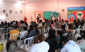 Um grupo de pessoas sentadas em um sala de aula, a frente duas mulheres palestram para estas pessoas.