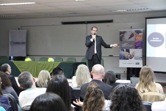 Na imagem um homem está no palco fazendo apresentação para os participantes que estão sentados na plateia.