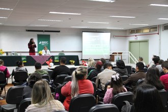 Na imagem duas professoras estão no palco em frente a plateia. Público observa pronunciamento de uma das docentes.