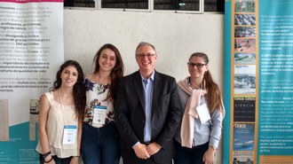 Quatro pessoas posam para foto em frente à banners de trabalhos científicos