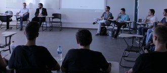 Várias pessoas em uma sala de aula, organizadas em círculo observam dois homens que falam sentados mais à frente
